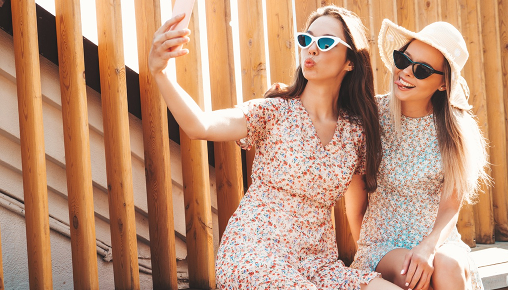 Two women in floral dresses taking a selfie against a wooden backdrop.