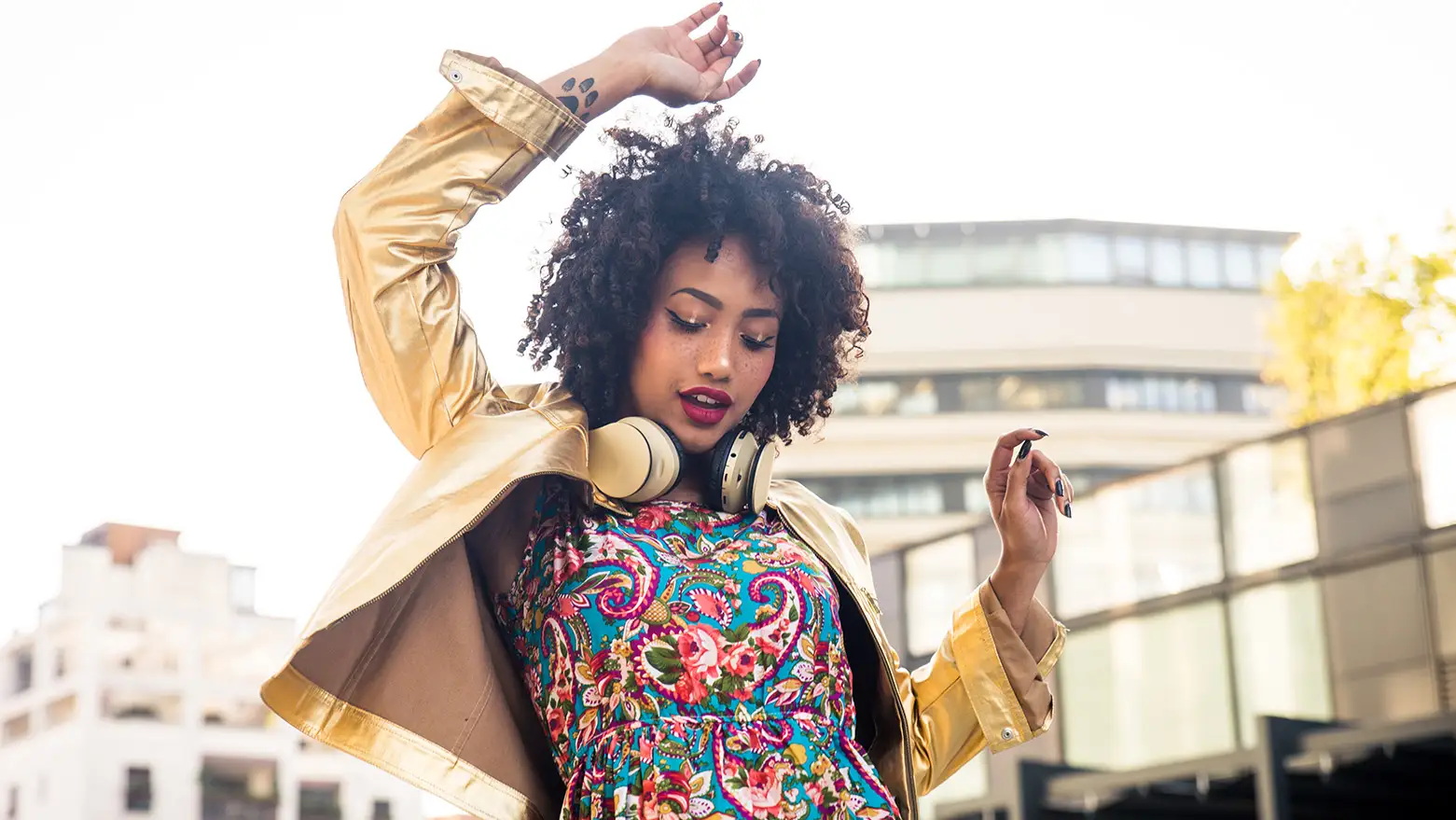 Stylish woman dancing outdoors in a colorful dress and gold jacket with headphones.