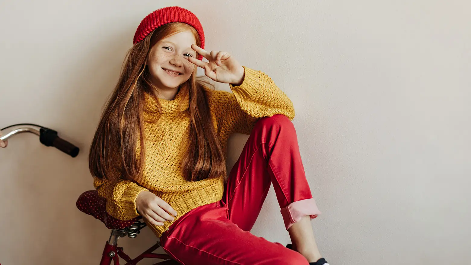 Smiling girl in a yellow sweater and red pants, posing playfully with peace sign.